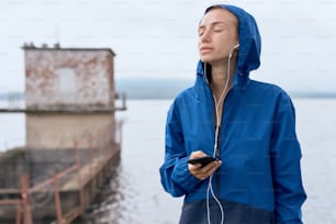 Retrato de mujer deportiva flaca de mediana edad con auriculares y teléfono inteligente al aire libre en verano, en un día sombrío con vista panorámica, con impermeable azul