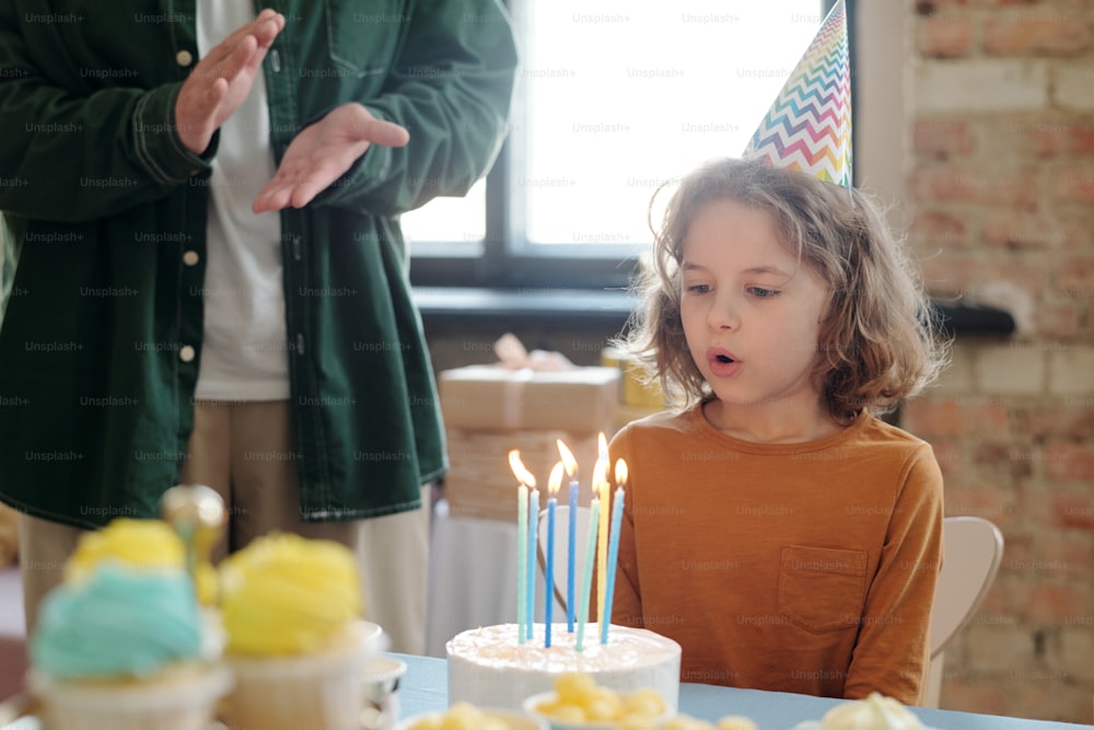 Little boy with long hair in party hat sitting at table with birthday cake and blowing candles on it