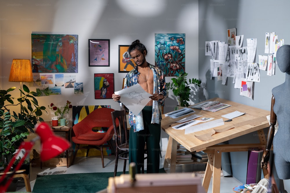 Moderne junge Modedesignerin, die in einem Raum steht, der mit Gemälden und Skizzen geschmückt ist und Muster für die Schneiderei betrachtet