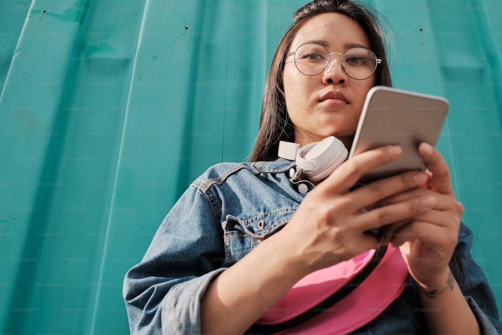 Below portrait of young asian student using smart phone outdoors, standing against cyan wall, wearing round glasses, blue jeans and pink belt bag