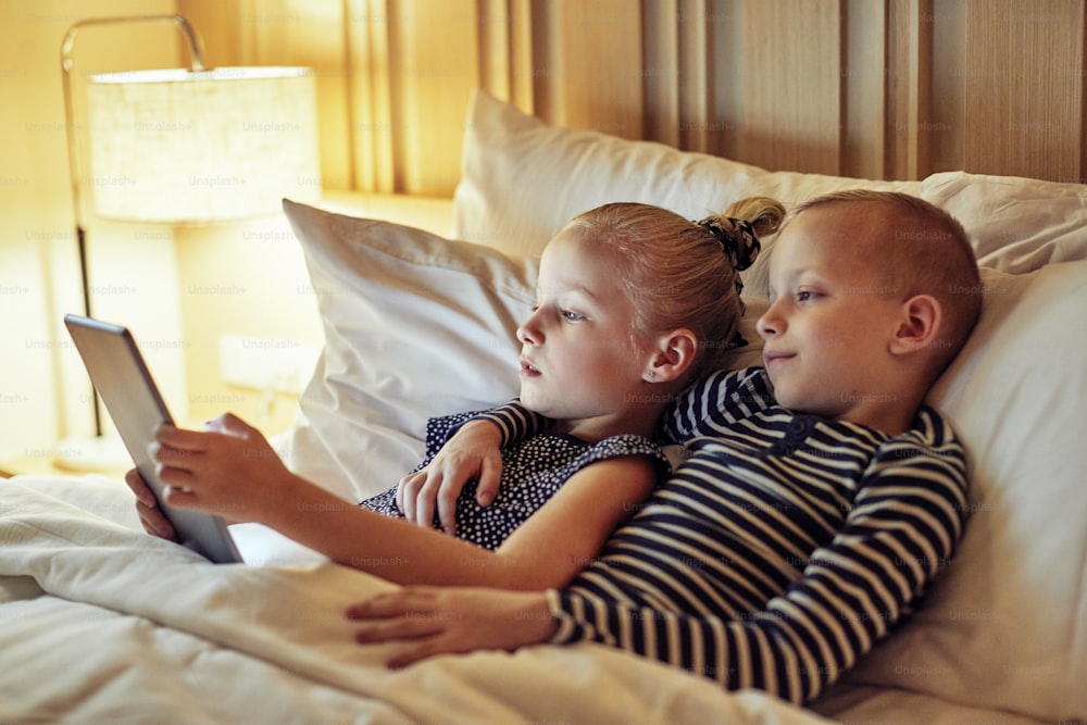 Cute little boy lying with his little sister in bed watching a video on a digital tablet before bedtime