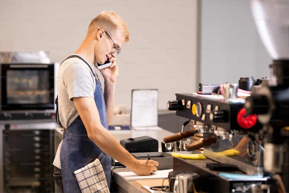 Foto Joven camarero o trabajador de la cafetería anotando los pedidos de  los clientes en el bloc de notas mientras habla con ellos por teléfono  móvil en el lugar de trabajo –