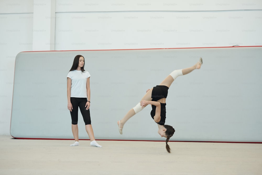 Junge Trainerin in Socken steht in der Sporthalle und bewertet die Fähigkeiten eines Cheerleading-Mädchens, das beim Training über den Kopf springt