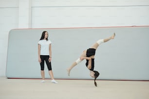 Giovane allenatore femminile in calzini in piedi nel palazzetto dello sport e valutando le abilità della ragazza cheerleader che salta sopra la testa durante l'allenamento