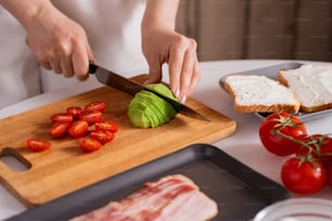 Hands of housewife with knife cutting fresh avocado and tomatoes on wooden board for sandwiches while cooking breakfast for herself