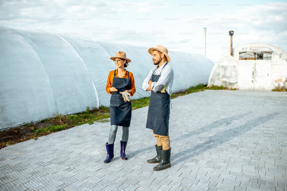 Due agricoltori o agronomi ben vestiti in piedi su una fattoria con serre sullo sfondo