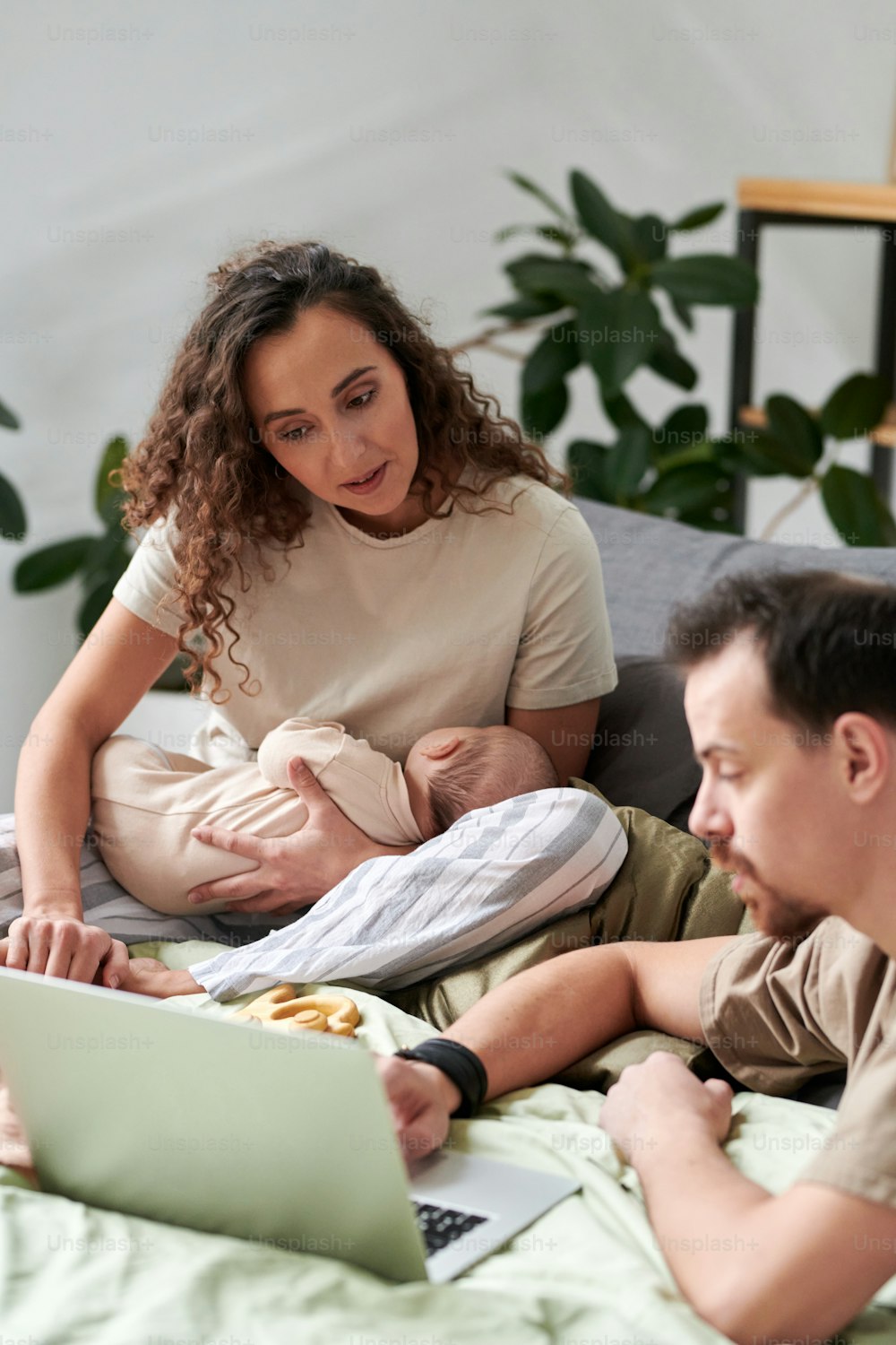 Young female with baby and her husband sitting on double bed and choosing new clothes for their son in online shop