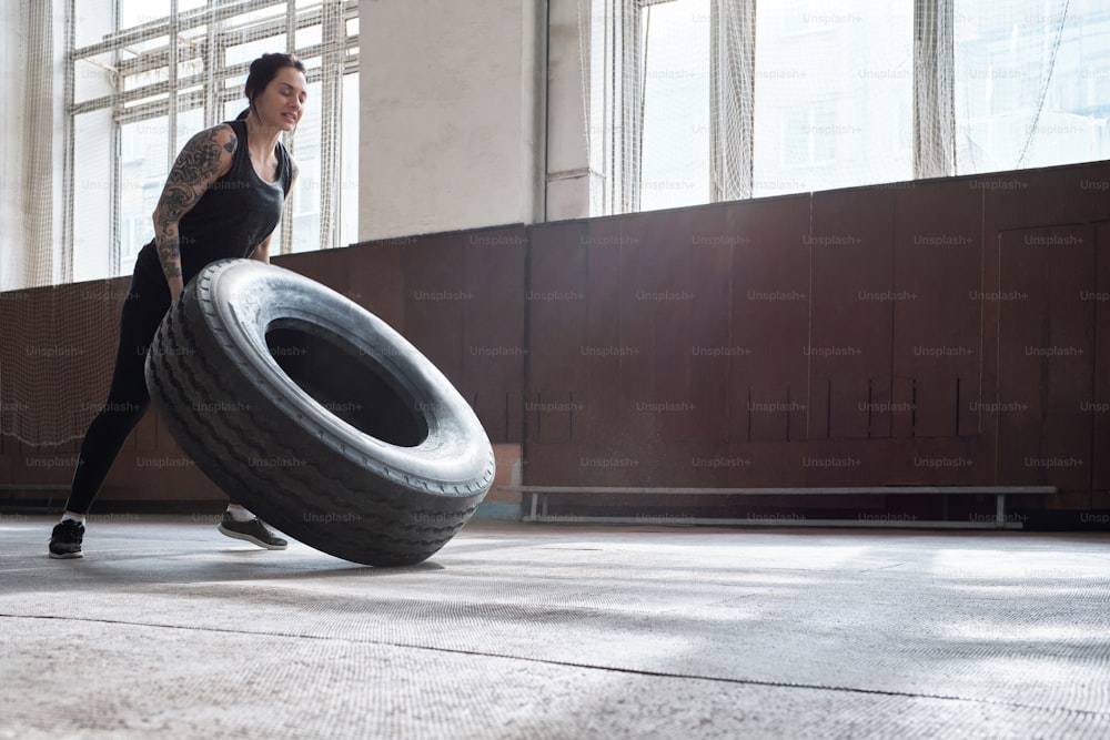 Entraînement croisé. Vue en contre-plongée d’une femme tatouée caucasienne athlétique renforçant le corps en retournant un pneu lourd dans une salle de sport