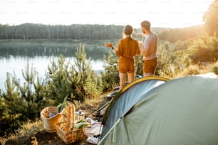 Young couple standing at the campsite, enjoying beautiful view on the lake, while traveling in the mountains on the sunset