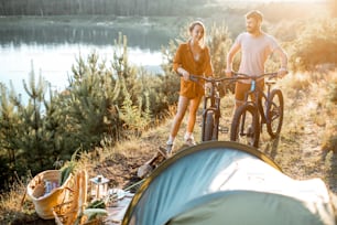 Young couple standing with mountain bicycles at the campsite, traveling in the forest near the lake on the sunset