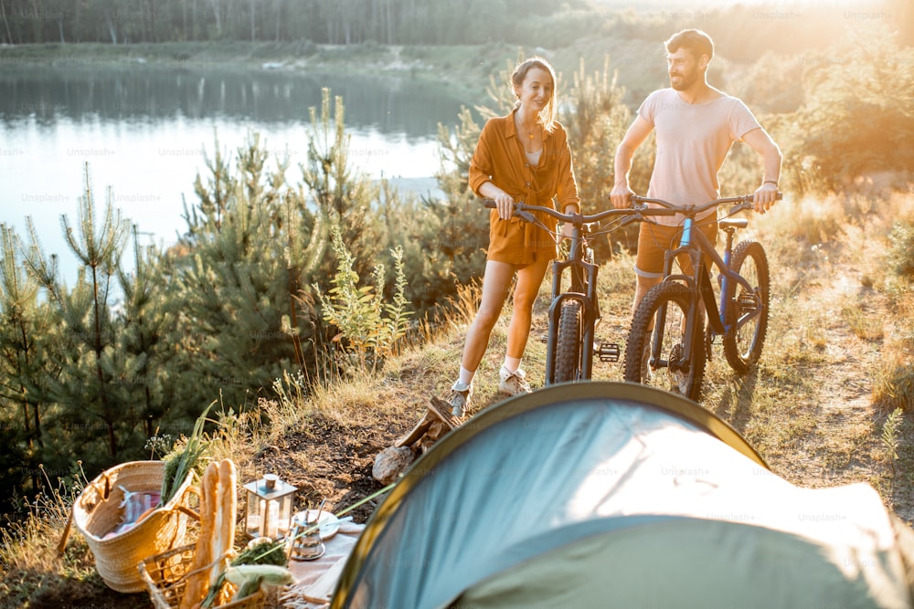 Jeune couple debout avec des vélos de montagne au camping, voyageant dans la forêt près du lac au coucher du soleil