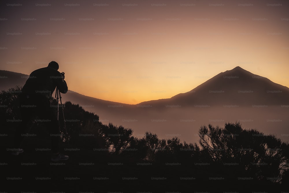 Fotografo in azione nel tramonto del paesaggio con alta montagna sullo sfondo e colori arancioni intorno alle persone attive selvagge che si godono l'attività di svago all'aperto