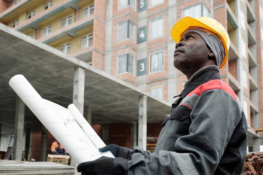 Ingeniero afroamericano en ropa de trabajo mirando hacia arriba en un condominio de gran altura mientras está de pie en el área de construcción contra el edificio