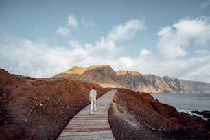 Woman walking on the picturesque wooden pathway through the rocky land with mountains on the background. Traveling on the north-west cape of Tenerife island, Spain