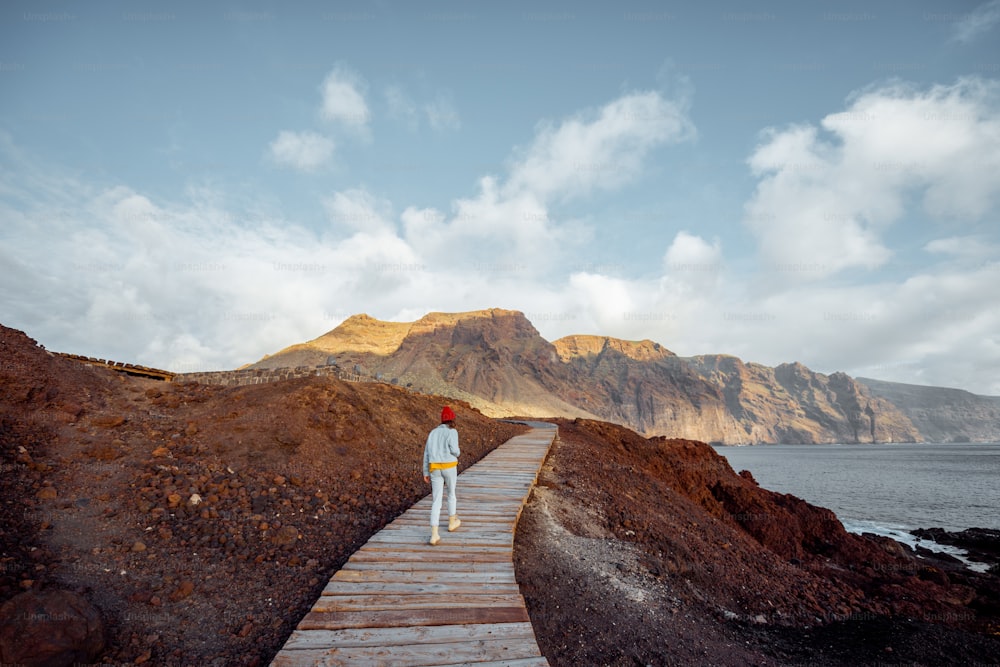 Woman walking on the picturesque wooden pathway through the rocky land with mountains on the background. Traveling on the north-west cape of Tenerife island, Spain