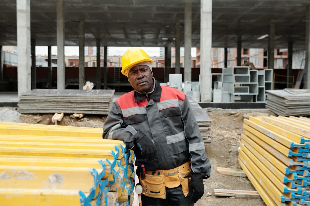Ernsthafter reifer schwarzer Mann in Arbeitskleidung und Schutzhelm schaut in die Kamera, während er neben einem Stapel gelber Baumaterialien steht