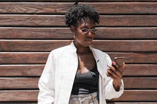 A portrait of a young charming black woman in oval sunglasses and white trench, and with curly Afro hair, using her cellphone while standing next to a striped wall made of wooden timbers