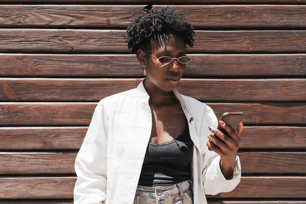A portrait of a young charming black woman in oval sunglasses and white trench, and with curly Afro hair, using her cellphone while standing next to a striped wall made of wooden timbers
