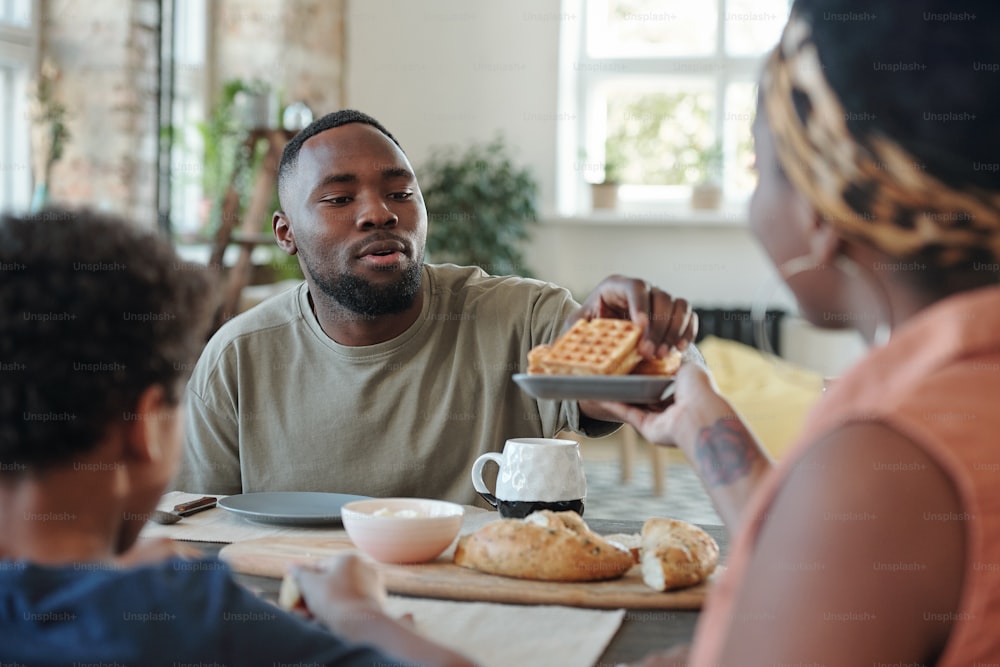 Joven africano sentado junto a la mesa de la cocina mientras toma un gofre casero del plato que sostiene su esposa durante el desayuno familiar por la mañana