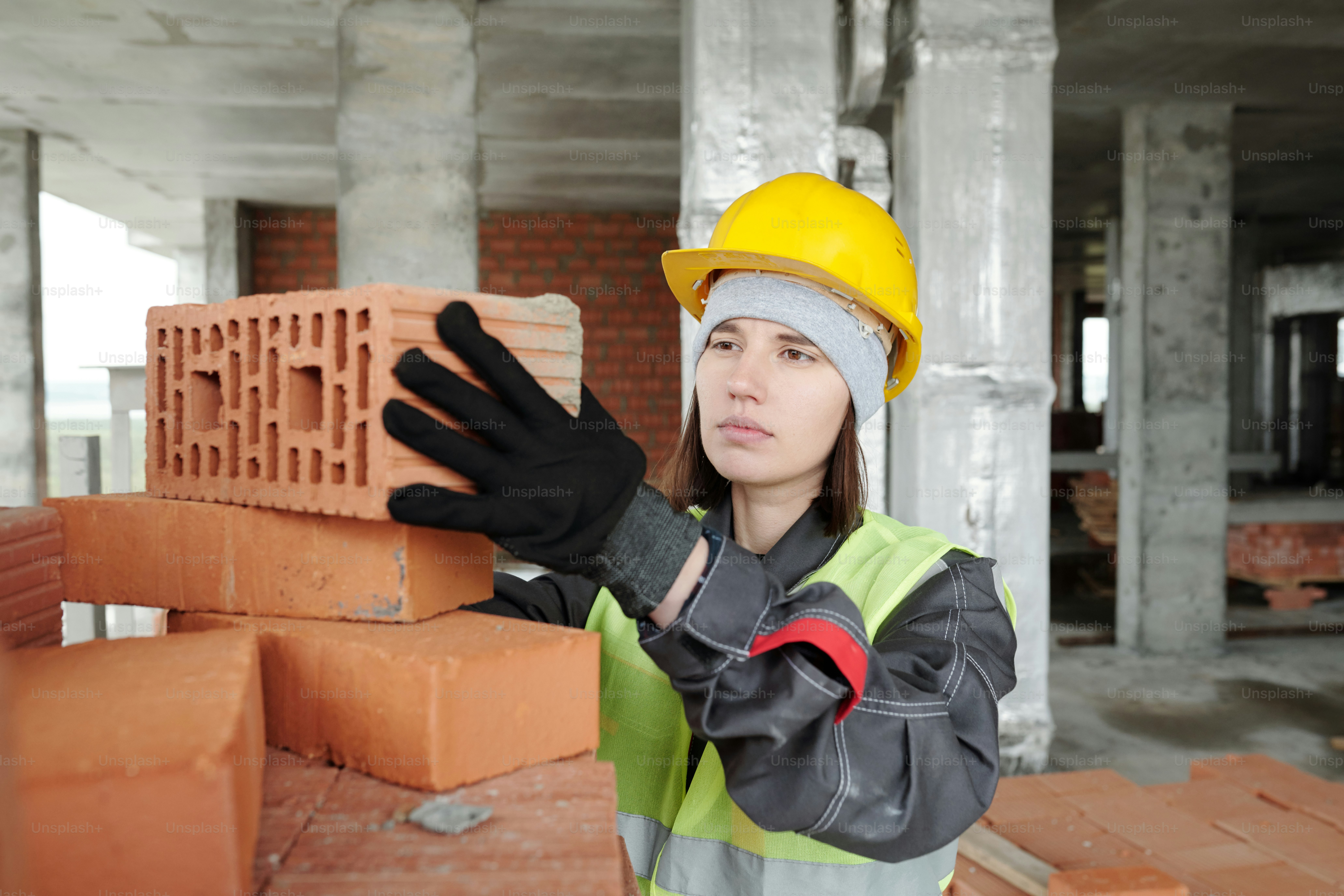 Giovane costruttore femminile serio in casco protettivo e orecchio da lavoro che prende il mattone dalla cima di un'enorme pila preparata per i lavori di costruzione