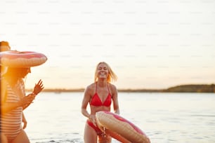Lachende junge blonde Frau im Bikini, die in einem See steht und einen Schwimmring hält und Spaß mit Freunden an einem See bei Sonnenuntergang hat