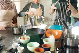 Joven entrenador de cocina masculino con delantal a rayas de pie junto al lugar de trabajo en la cocina y explicando al grupo de alumnos cómo cocinar comida sabrosa