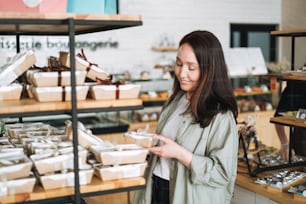 Adult smiling brunette woman forty years with long hair in stylish shirt choosing sweets in cafe, woman having shopping in confectionery