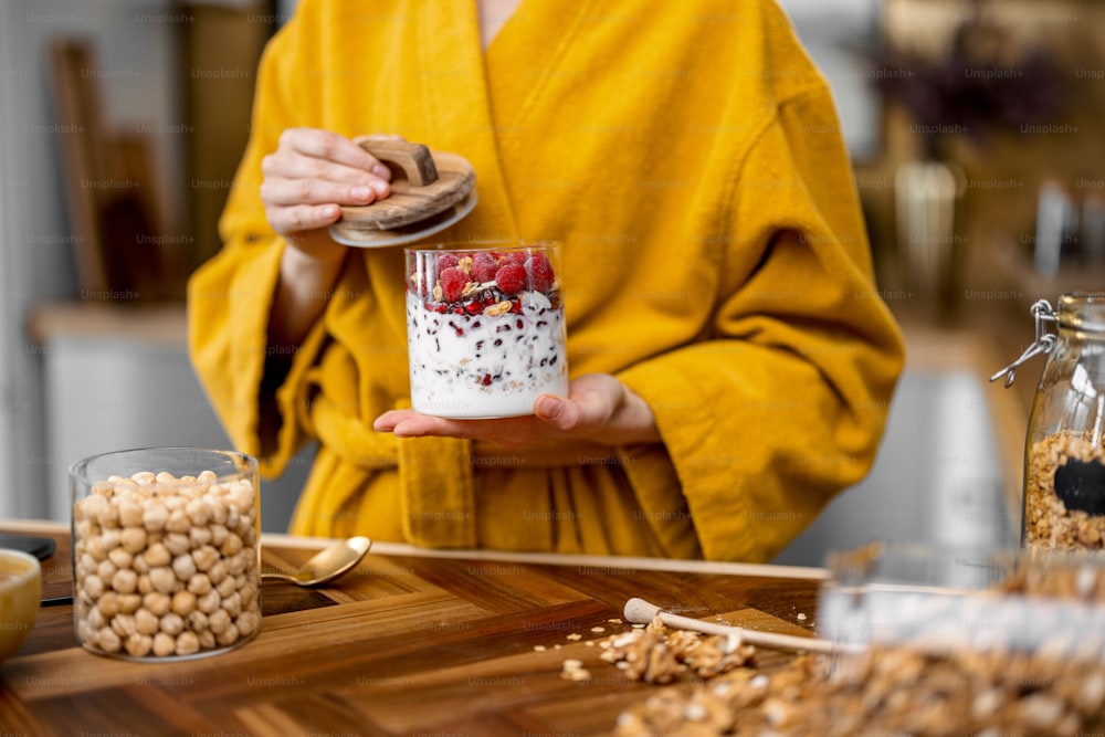 Mulher nova em roupão amarelo desfruta de café da manhã saudável de cereais com iogurte e bagas na tigela na cozinha em casa pela manhã