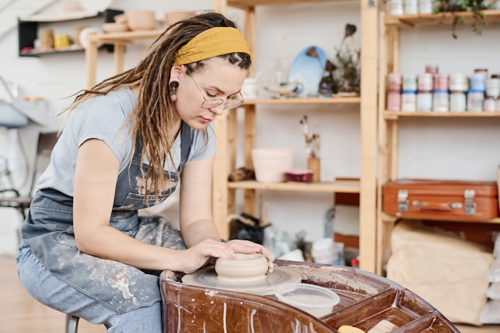 Giovane donna creativa in abbigliamento casual e grembiule che si piega sul tornio di ceramica e crea un nuovo oggetto di terracotta in officina