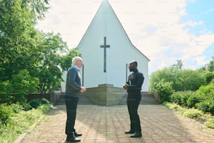 Seitenansicht eines jungen Pastors und eines älteren männlichen Gemeindemitglieds, die auf dem Kirchhof diskutieren, während sie voreinander stehen