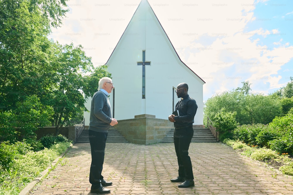 Vista lateral do jovem pastor e do paroquiano masculino sênior discutindo no pátio da igreja enquanto estão um na frente do outro