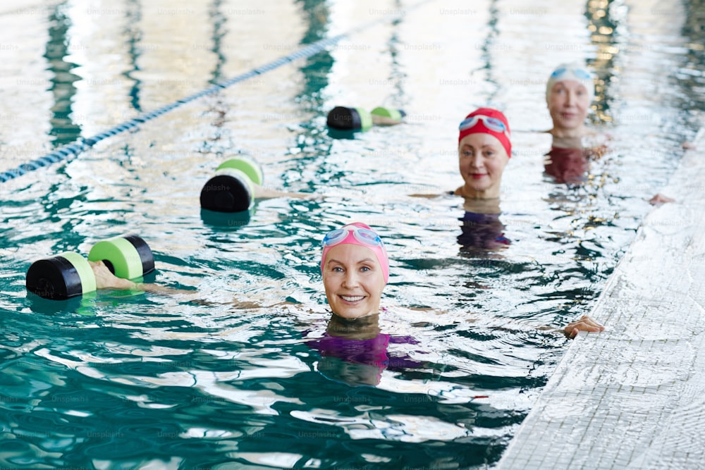 Fila de mujeres mayores felices en traje de baño estirando sus brazos derechos mientras levantan pesas durante el entrenamiento en el agua