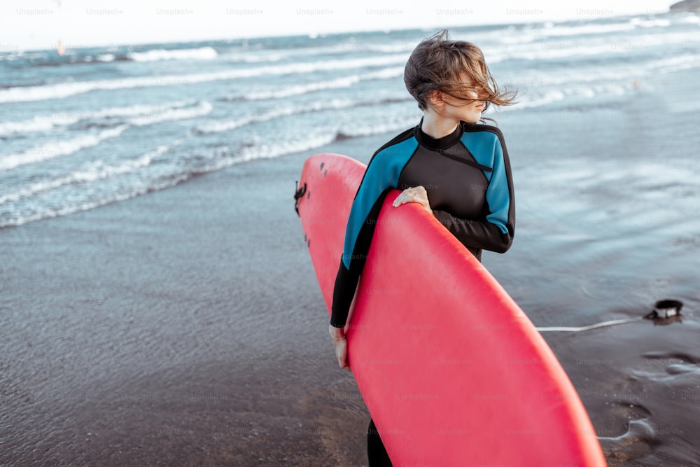 Porträt einer jungen Surferin im Badeanzug, die mit rotem Surfbrett am Strand steht. Aktiver Lifestyle und Surfkonzept