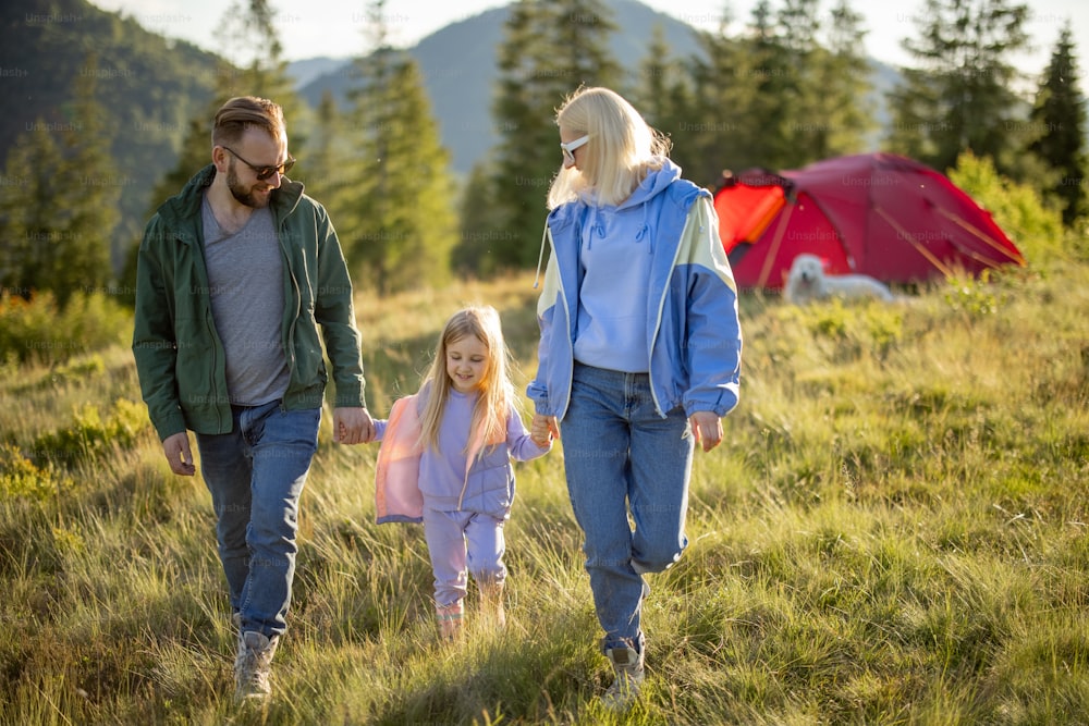 Young caucasian family with little girl walk together on green meadow while traveling with tent in the mountains. Happy family summer vacation at campsite