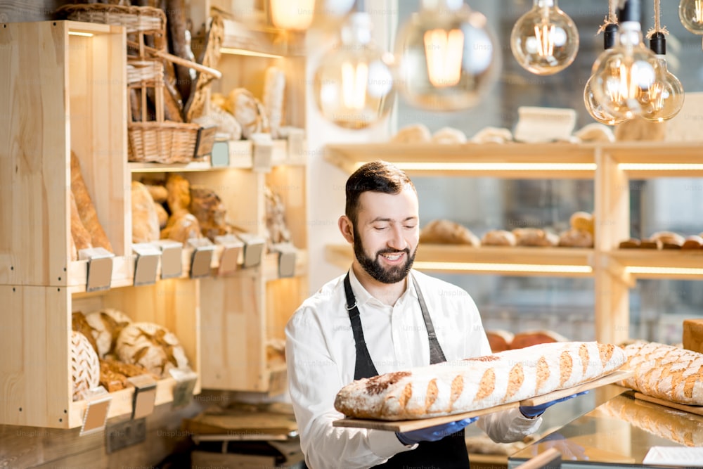 Handsome seller in uniform holding a big loaf of bread in the store with bakery products