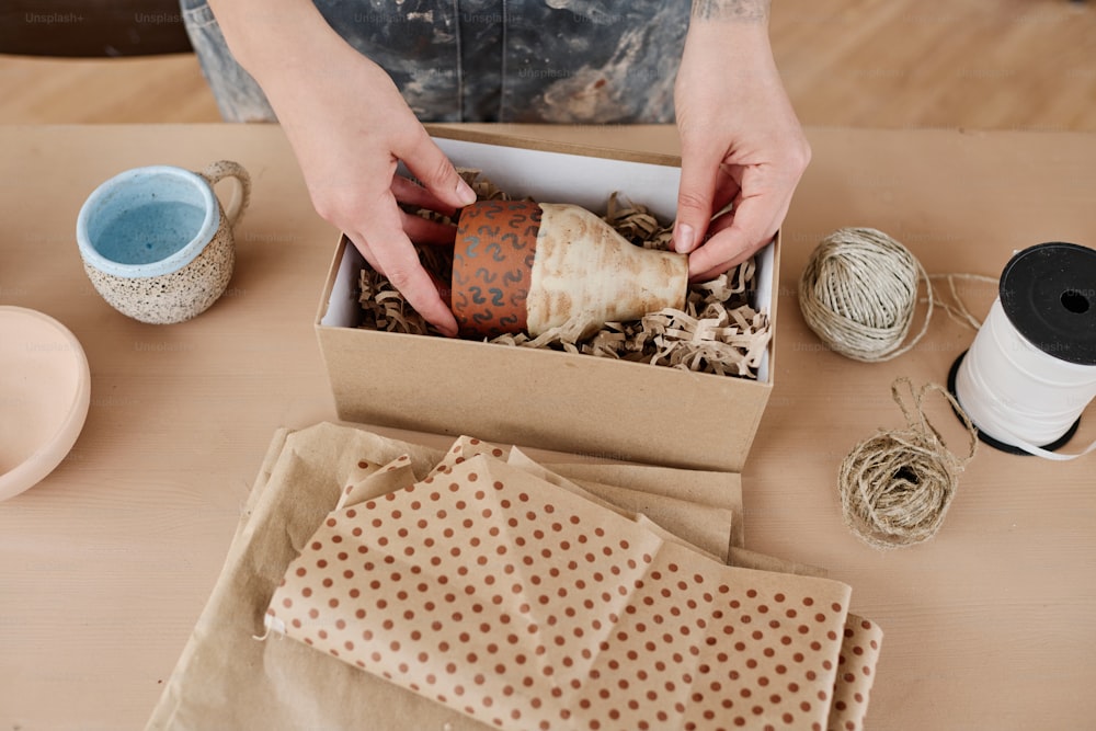 Über dem Winkel der Hände der Handwerkerin, die handgefertigte Kanne oder Vase in die Box steckt, während sie einen der handgefertigten Artikel für den Online-Kunden verpackt
