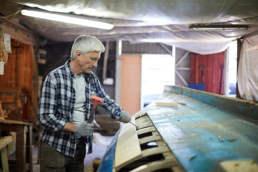 Maestro envejecido con cincel y martillo de pie junto a la construcción de la vasija y el procesamiento de sus tablones