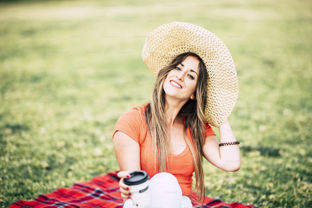 Cheerful young woman with hat and long blonde hair enjoy outdoor leisure activity sit down on the grass - people with happy life enjoy the nature and relax time