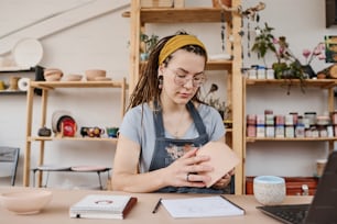 Joven empresaria creativa en ropa casual sosteniendo un artículo de loza mientras evalúa su calidad durante el trabajo en una pequeña tienda o estudio