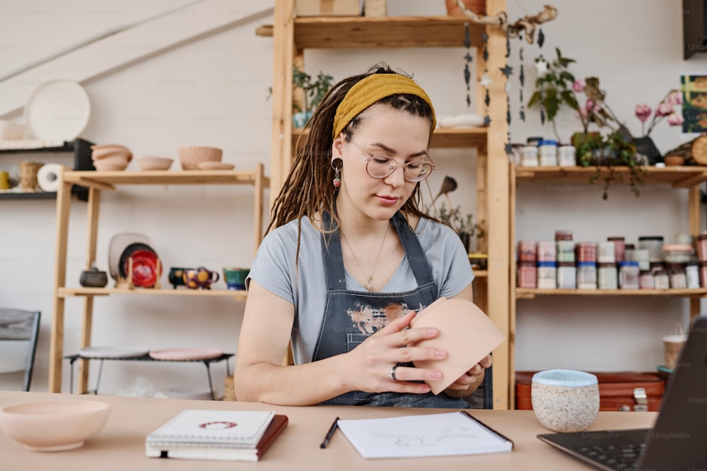 Young creative businesswoman in casualwear holding earthenware item while evaluating its quality during work in small shop or studio