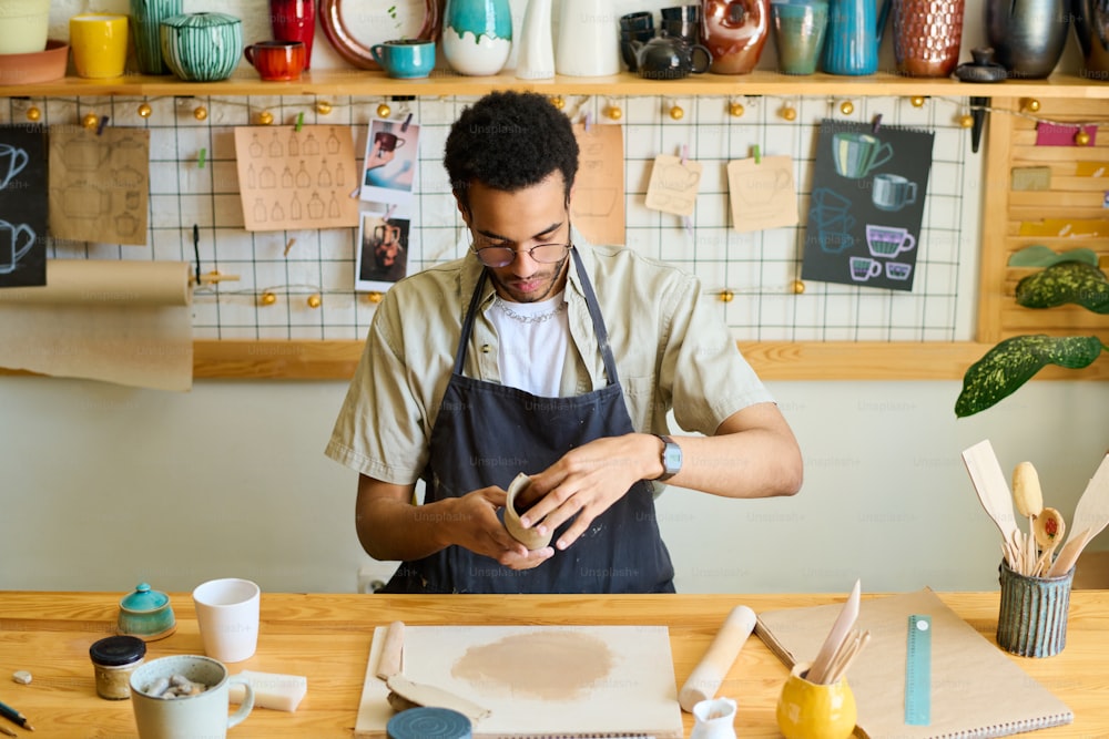 Junger kreativer schwarzer Mann in Arbeitskleidung, der neue Tonartikel zum Verkauf kreiert, während er in der Werkstatt am Tisch gegen das Regal mit Steingut sitzt