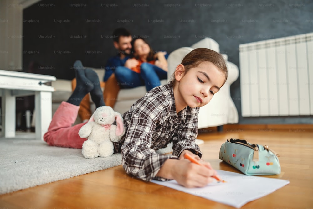 Child lying down on stomach on the floor drawing. In blurry background parents hugging. Happy family.