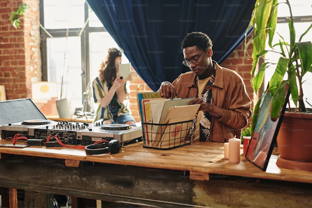 Joven afroamericano mirando a través de su colección de discos de vinilo en caja de pie sobre la mesa junto a la cabina de DJ y los auriculares