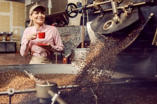 A coffee factory worker drinking cup of coffee next to a roasting machine in facility.