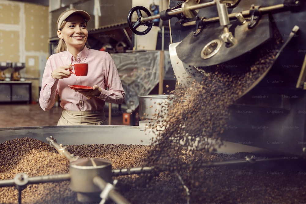 A coffee factory worker drinking cup of coffee next to a roasting machine in facility.