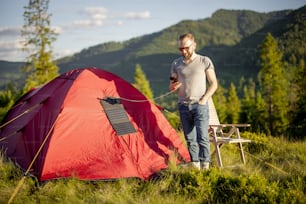Man uses phone and charges it from portable solar panel while traveling with tent in the mountains. Concept of modern technologies for hiking and renewable energy