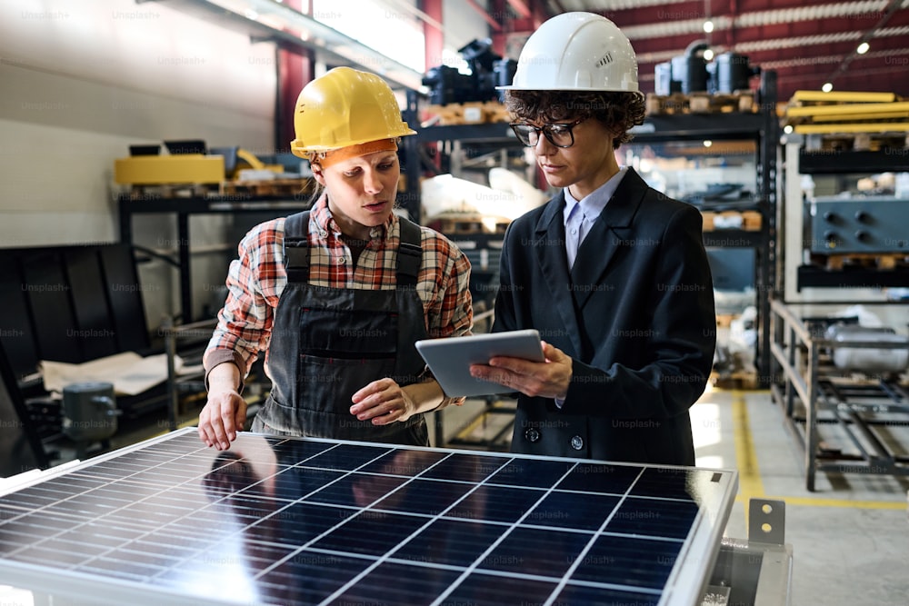 Gerente confiado con tableta que muestra datos en línea de una joven ingeniera sobre las características de los paneles solares producidos en la fábrica
