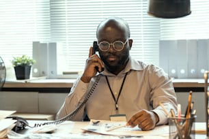 Serious African American FBI agent in formalwear speaking on telephone while sitting by workplace and looking through documents