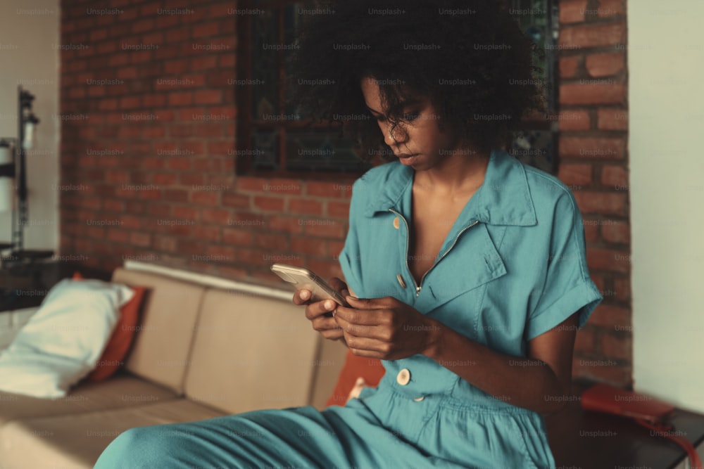 A portrait of a cute young Hispanic woman in blue overalls sending a message to her friend while sitting in a guest-room of her brick house, on a sofa; a copy space place on the left for an ad message