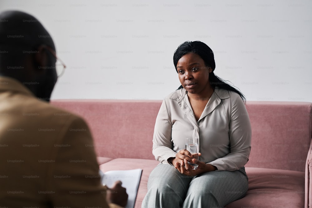 Young woman with glass of water sitting on couch and looking at counselor while listening to his advice during psychological session